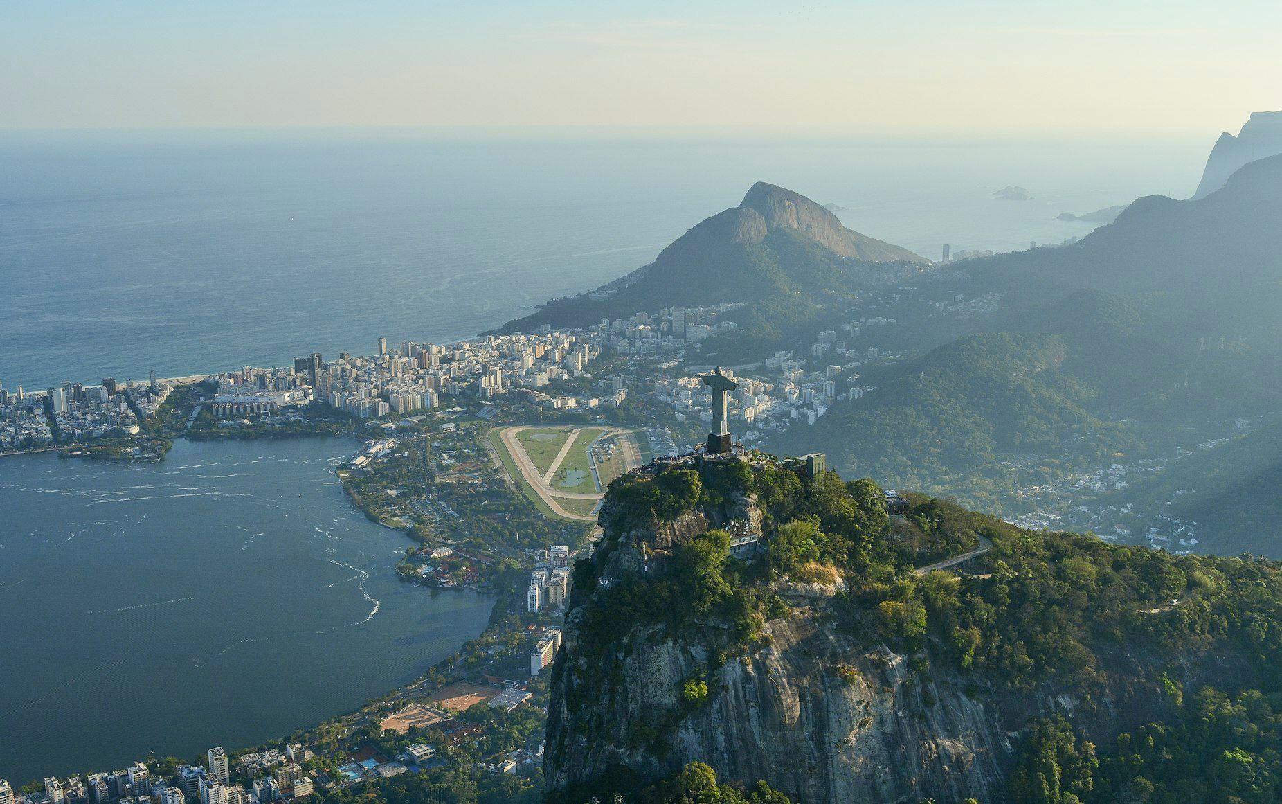 An aerial view of the Christ the Redeemer statue on a mountain, overlooking a coastal city, a lagoon, and the ocean at sunrise.