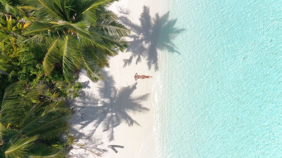 An aerial view of a person lying on a white sand beach by turquoise water, under the shadows of palm trees.