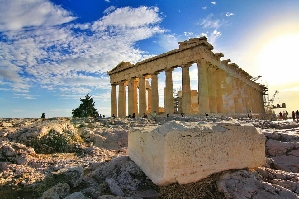 An ancient Greek temple with large columns stands on a rocky hill under a blue sky with white clouds at sunset.