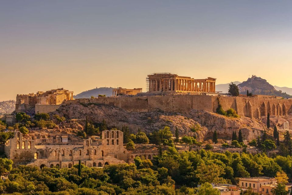 Ancient Greek temples and ruins sit atop a rocky hill overlooking a dense forest during a golden sunset.