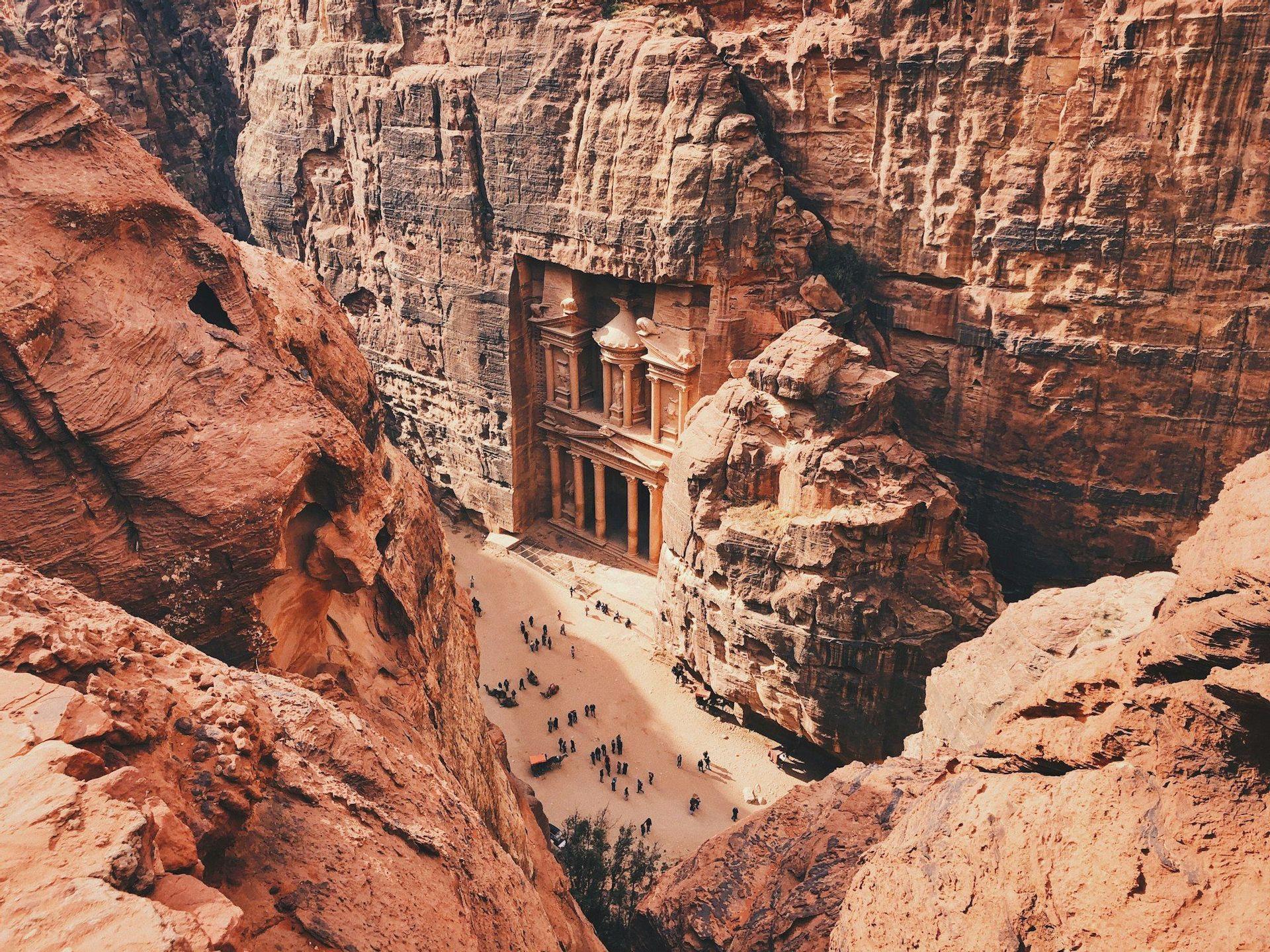 An ancient temple facade carved directly into a red sandstone cliff, seen from a high vantage point with people below.