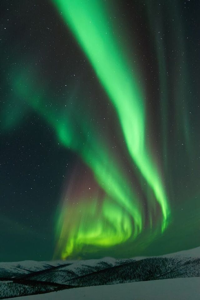 Bright green bands of the aurora borealis glow in the starry night sky above a snowy mountain range.