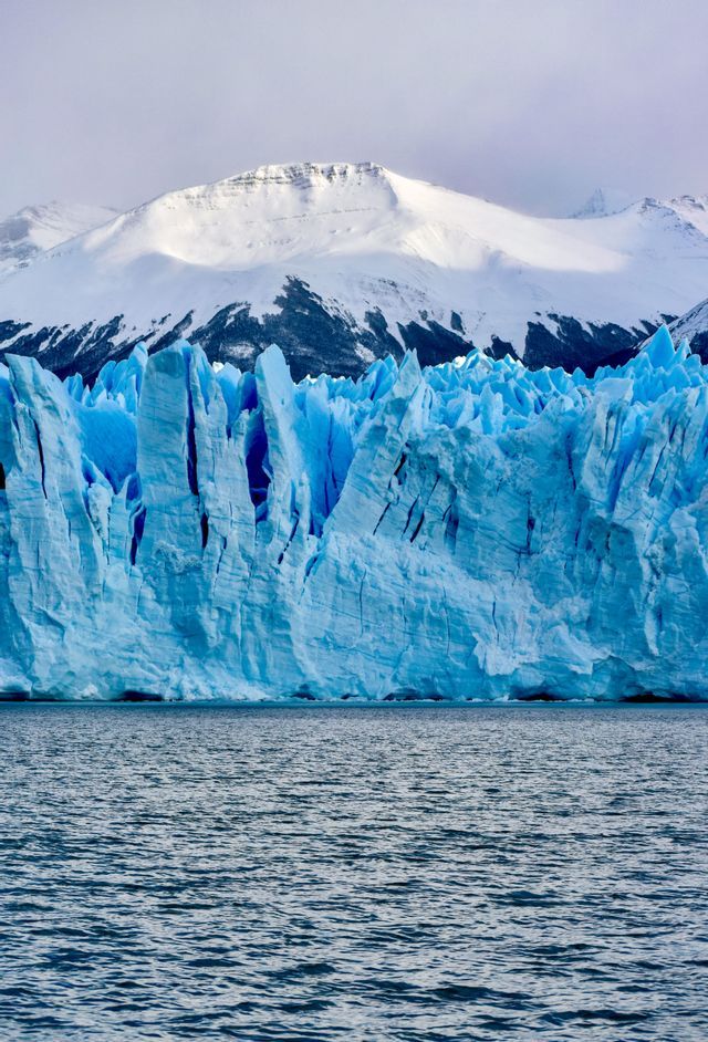 A massive blue glacier rises from the dark water, with snow-covered mountains in the background.