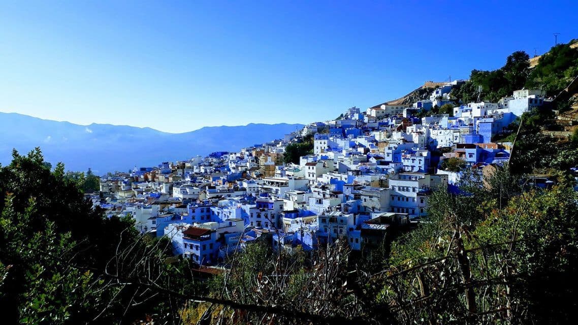 A city of blue and white buildings covers a green hillside, with distant mountains under a clear sky.