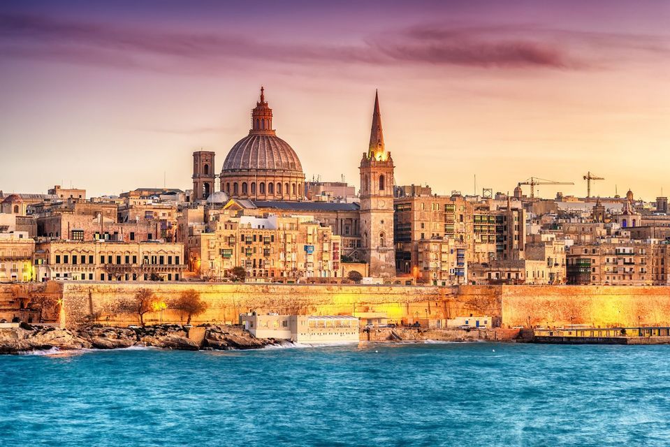 A coastal city with a large dome and steeple is lit up along the water's edge under a colorful sunset sky.