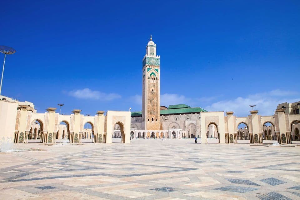 A grand mosque with a tall, ornate minaret stands behind a large, patterned plaza under a bright blue sky.