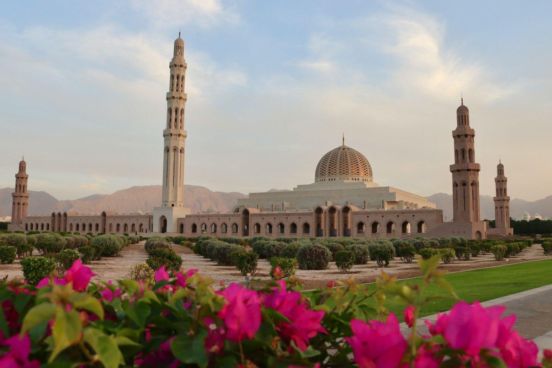 A large mosque with a central dome and several minarets, viewed over a landscaped garden with pink flowers in the foreground.