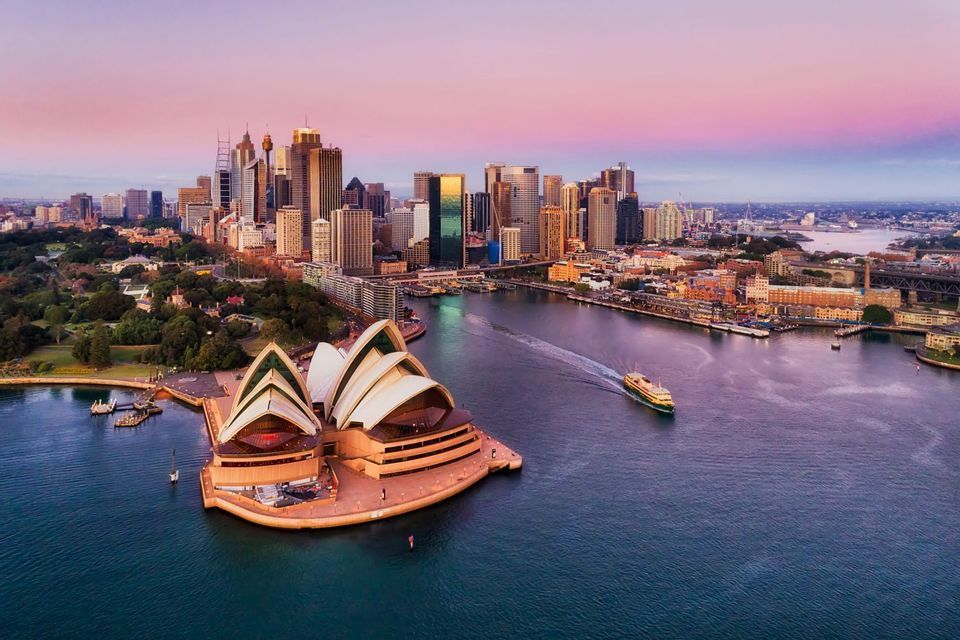 An aerial view of an iconic opera house and a city skyline at sunset, with a ferry crossing the harbor.