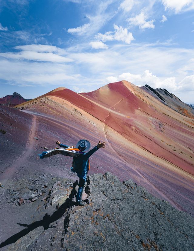 A person in a hat stands on a rocky outcrop with arms outstretched, overlooking a range of colorful, striped mountains under a blue sky.