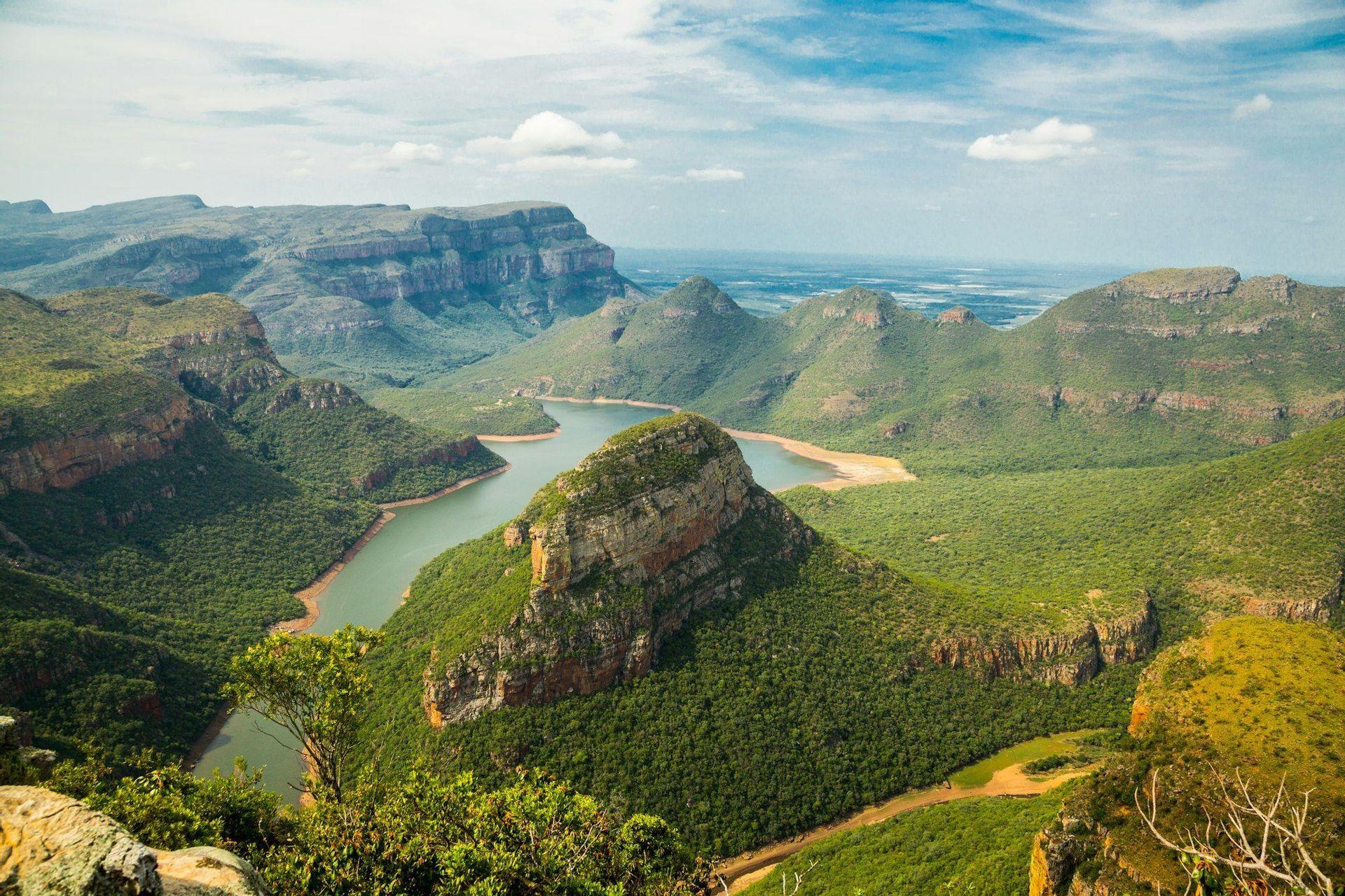 A panoramic view of a river winding through a vast green canyon with large, flat-topped mountains under a blue sky.