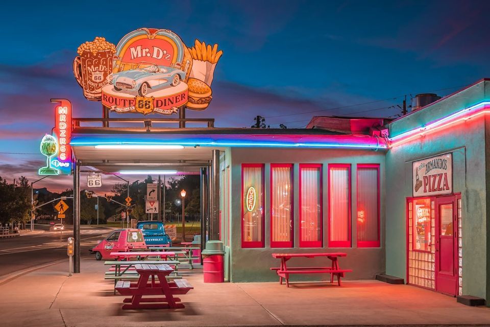 The exterior of Mr. D'z, a retro Route 66 diner, glows with colorful neon signs at twilight.