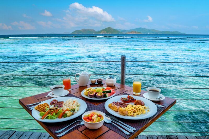A full tropical breakfast spread for two served on a wooden table overlooking turquoise ocean waters.