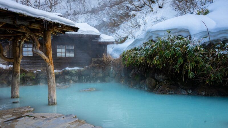 Traditional snowy onsen with milky blue water and rustic wooden building.