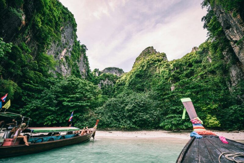Two traditional longtail boats anchored on a pristine beach, surrounded by lush green cliffs and dense jungle vegetation in a secluded cove in Thailand.