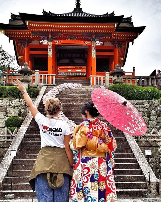 A WeRoad group leader stands with a woman in a traditional kimono and parasol at the base of stairs leading to an orange temple.