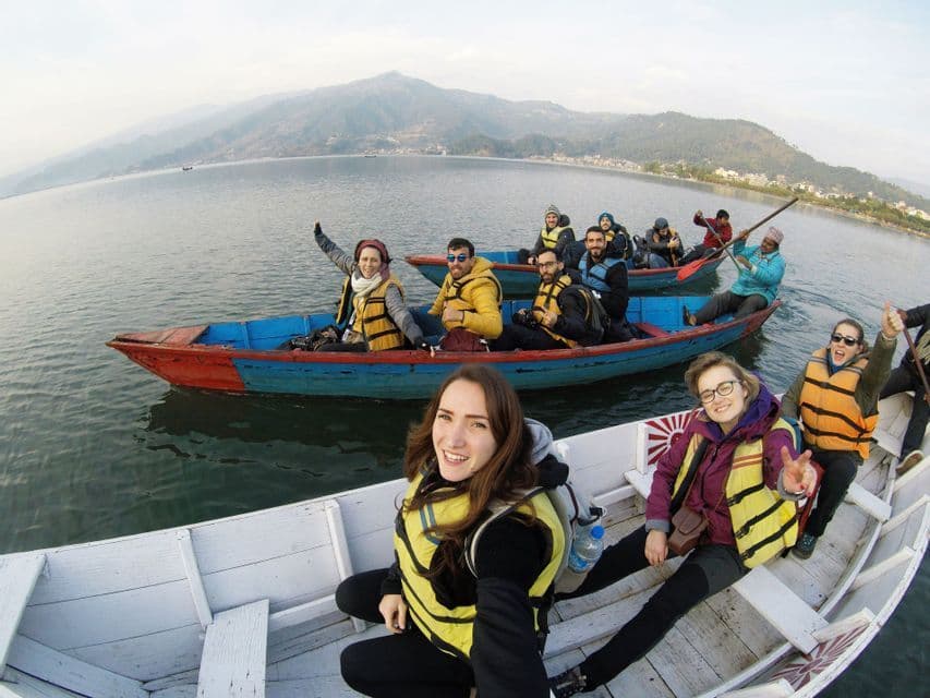 A WeRoad group trip smiles for a selfie while riding in colorful boats on a large lake, with hazy mountains in the background.