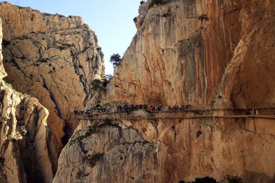 A WeRoad group trip walks single-file along a narrow boardwalk attached to the sheer rock face of a large canyon.