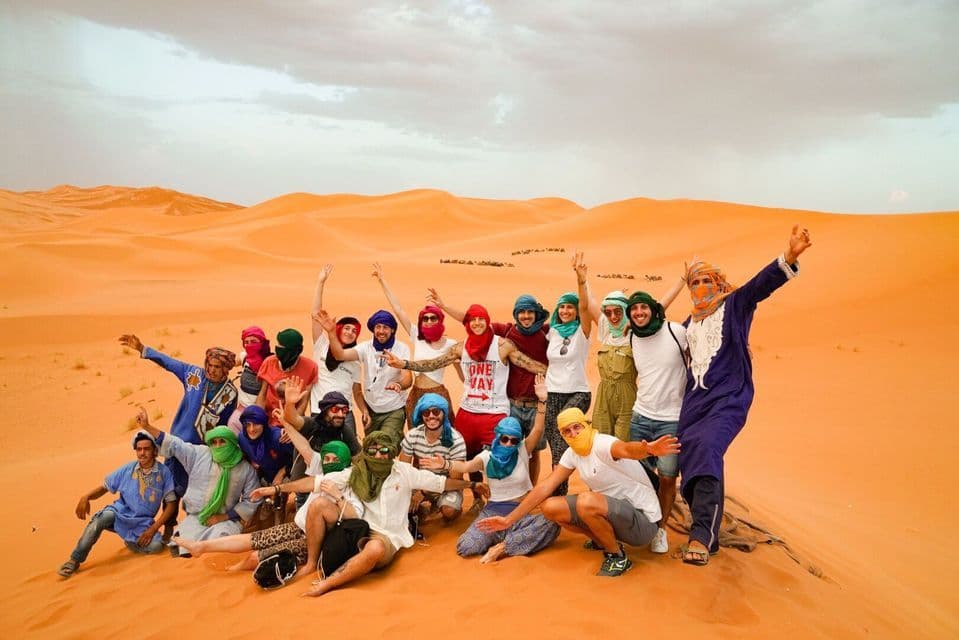 A large WeRoad group trip with colorful headscarves posing together for a photo on vast orange sand dunes.