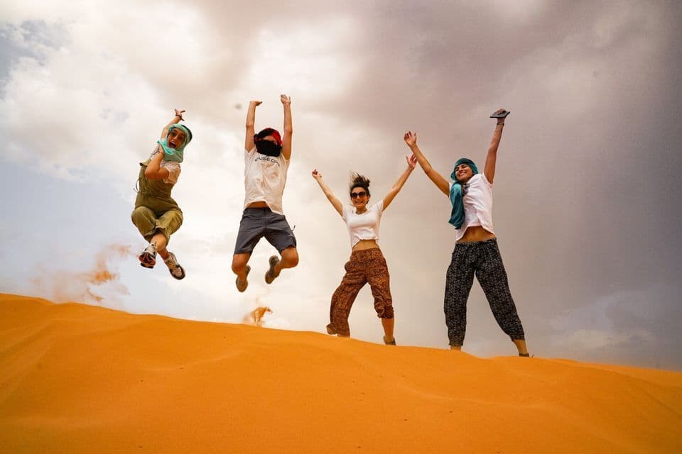 A WeRoad group trip of four people jumping in the air on an orange sand dune under a cloudy sky.