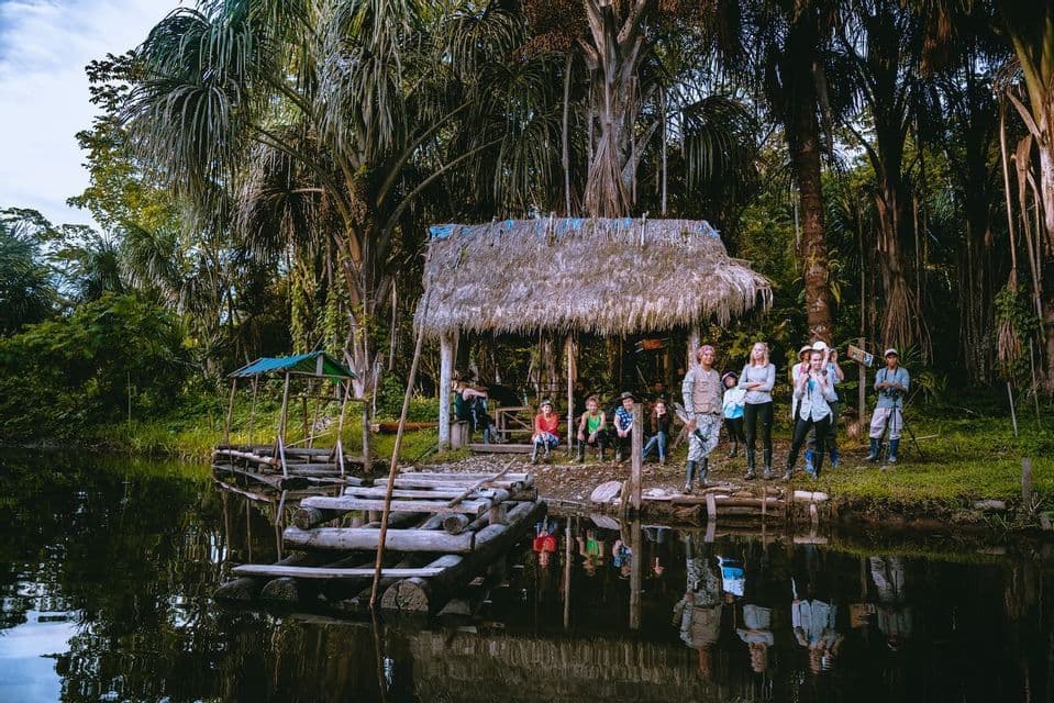 A WeRoad group trip stands on the bank of a calm river in the jungle, next to a wooden dock and a hut with a thatched roof.