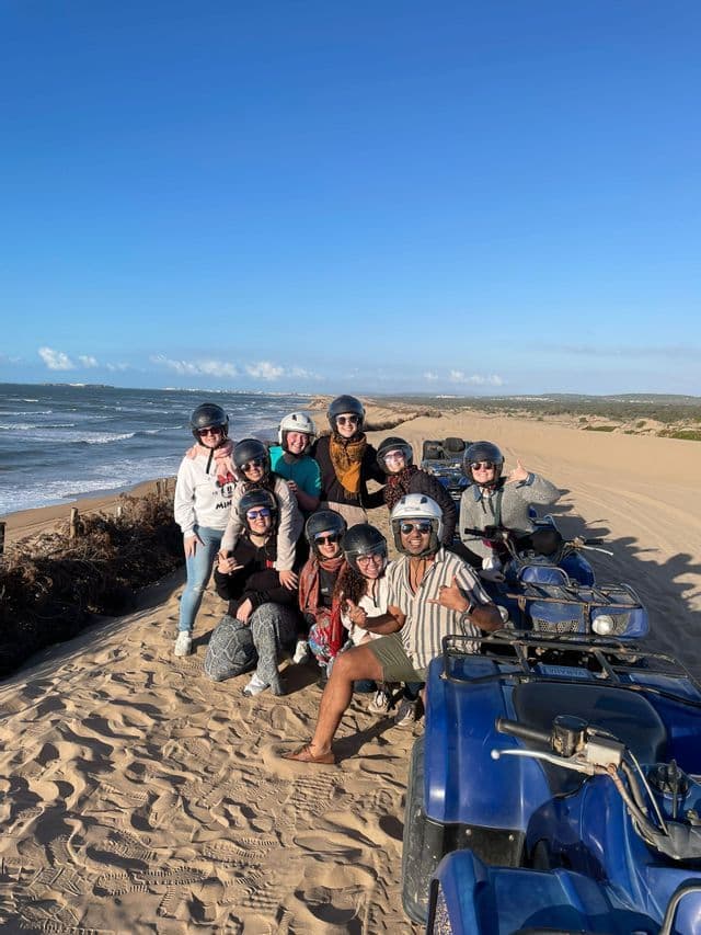 A WeRoad group trip wearing helmets poses for a photo on sand dunes with their quad bikes, with the ocean in the background.