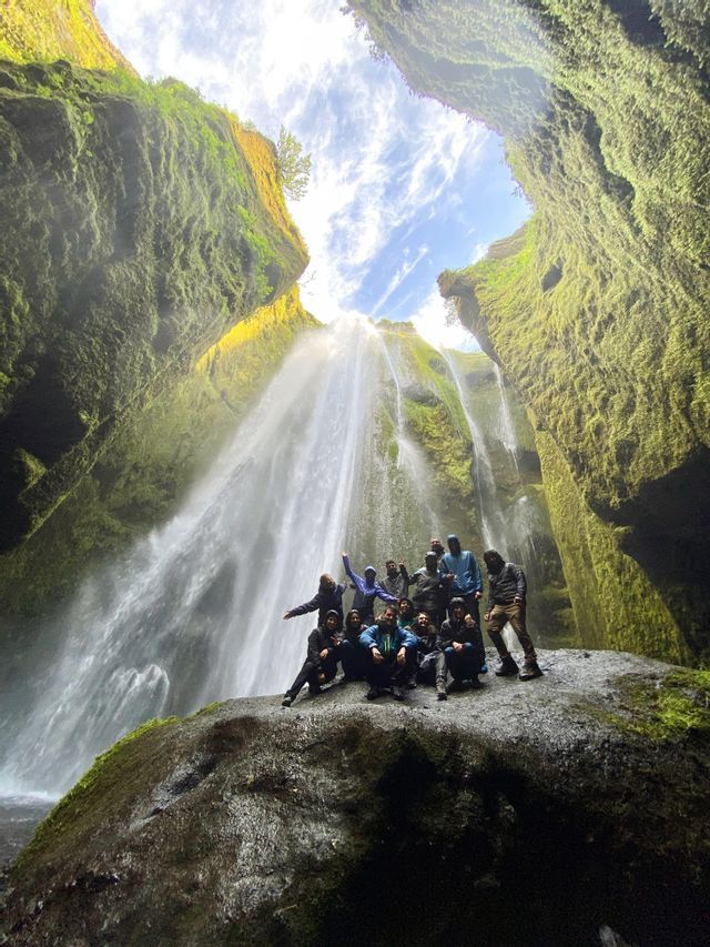 A WeRoad group trip poses on a large rock at the base of a waterfall inside a mossy canyon.
