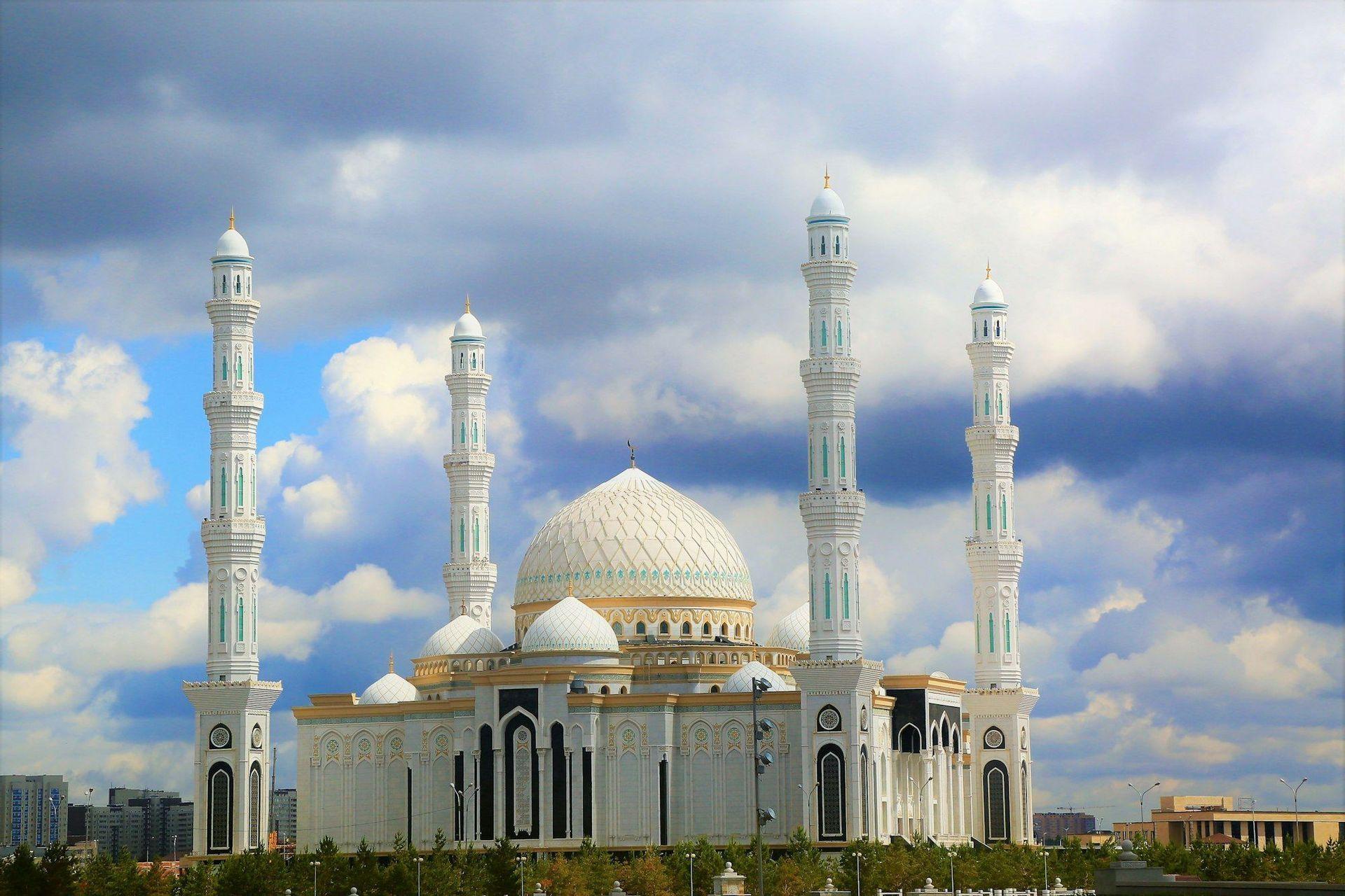 A large, ornate white mosque with domes and tall minarets stands against a backdrop of dramatic blue and gray clouds.