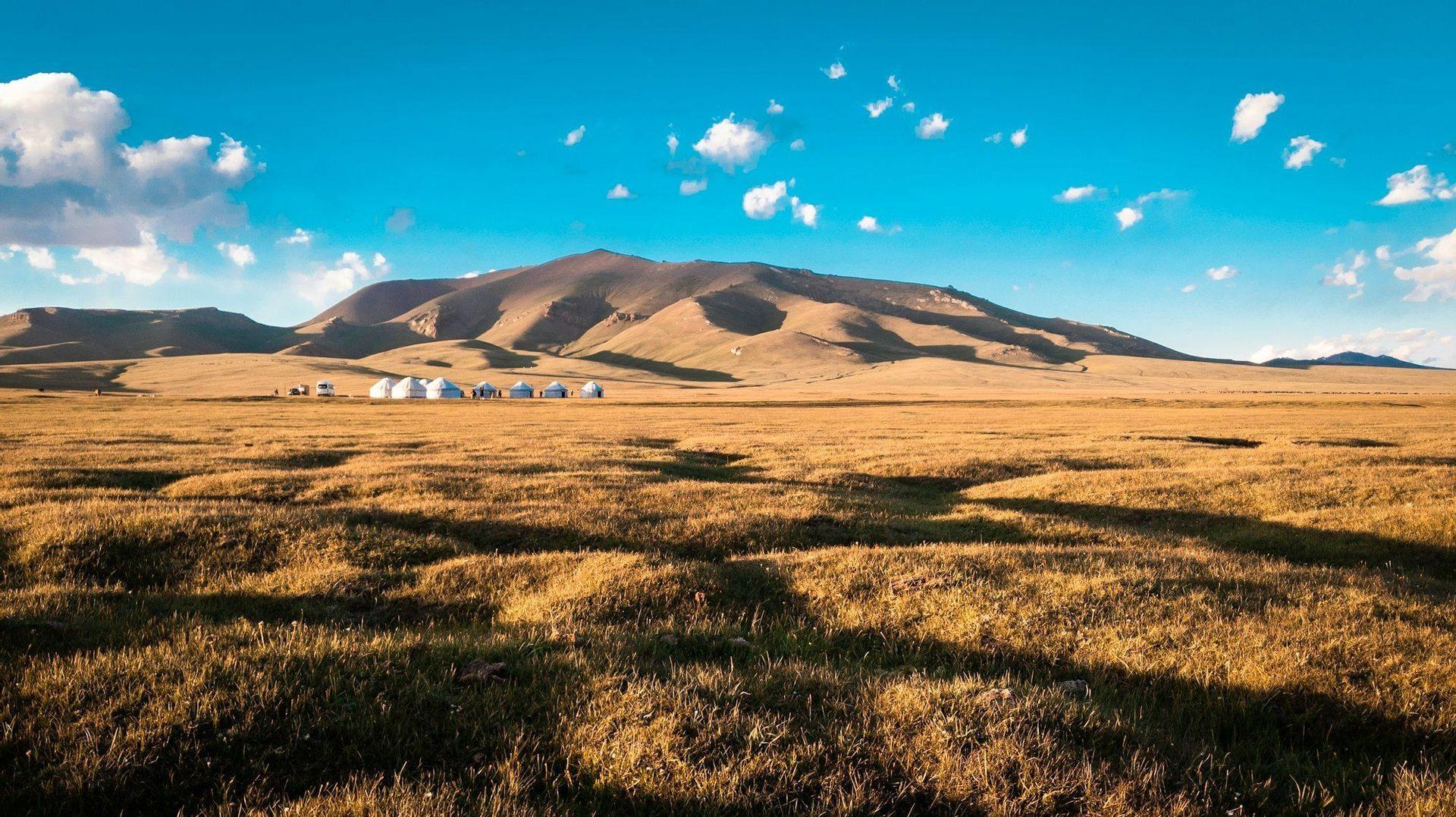 A group of white yurts are set up on a vast grassy plain at the base of a large mountain under a blue sky with scattered clouds.